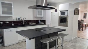 A modern kitchen with a central island, stainless steel appliances, and a dining area in the background. - Domaine la Griffe - Maison Mitoyenne Cou (La Roche-sur-Foron)