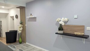 A modern reception area with a grey wall, a wooden bench, a small shelf with a vase of white flowers, and a black chair. - Domaine la Griffe - Maison Mitoyenne Cou (La Roche-sur-Foron)