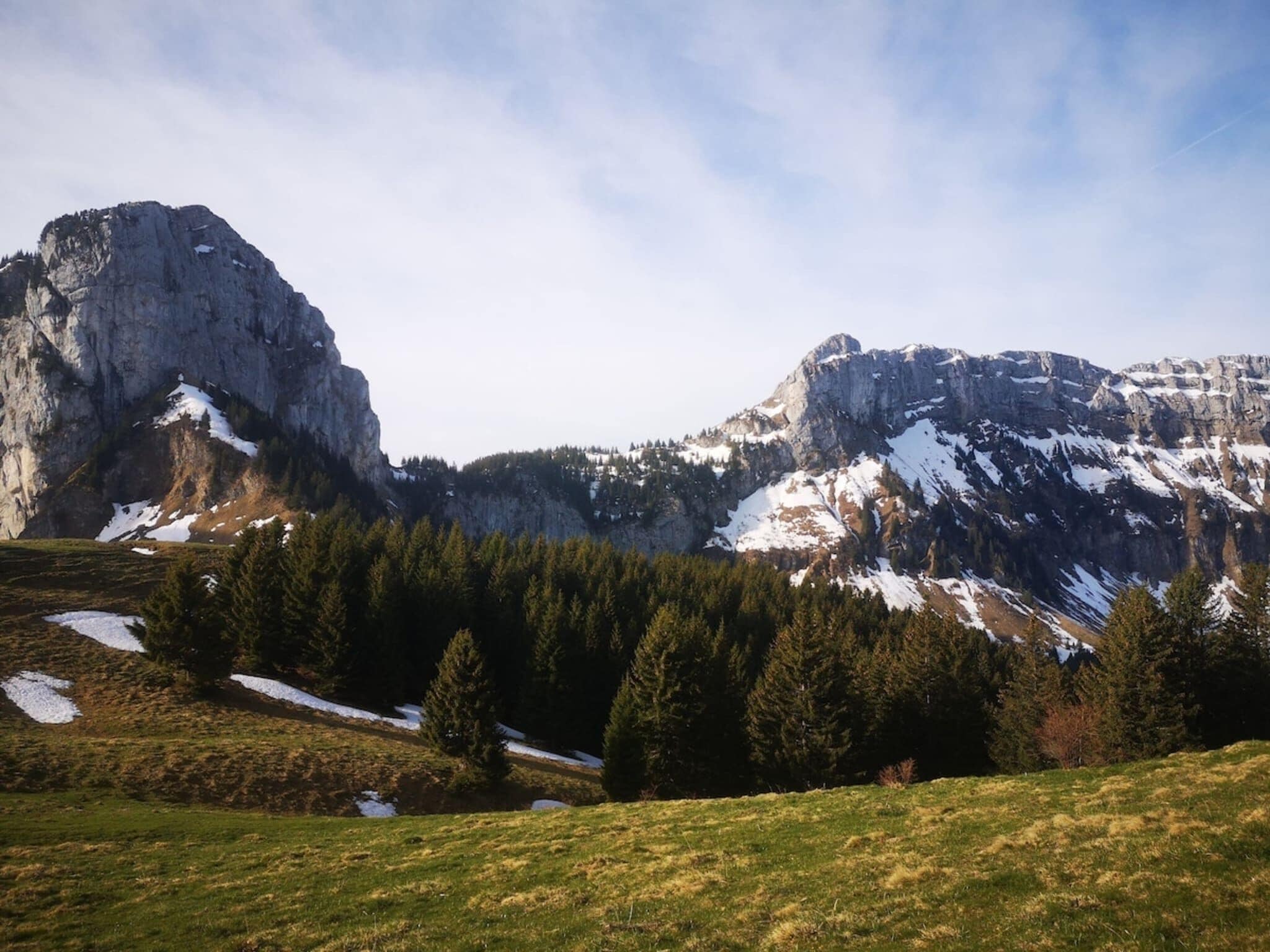 A mountainous landscape with a prominent rocky peak, dense forest, and patches of snow.