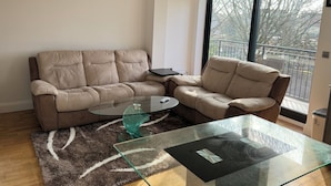 Ein modernes Wohnzimmer mit einem beige farbenen Sofa, einem Glastisch und Blick auf einen Balkon mit Geländer.