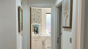 Ein Flur, der zu einem Badezimmer mit Toilette, Waschbecken und Dusche führt. An den Wänden hängen Bilder in Rahmen und auf dem Boden liegt ein Teppich.