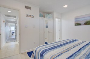 A bedroom with a striped bedspread, white walls, a painting of a beach, and a hallway leading to another room. - Duck Tales - Oceanfront : Steps from the beach : Newly renovated! (Kitty Hawk)