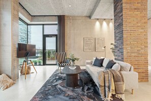 Ein modernes Wohnzimmer mit einer Couch, einem Couchtisch, einem Fernseher auf einem Stativ und einem großen Fenster mit Blick auf Gebäude.