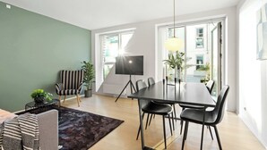 A modern living room with a sofa, a striped armchair, a dining table with chairs, a TV on a stand, and a large window with a view of buildings. - The Iceberg Residence (Aarhus)