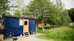 A small blue cabin with a wooden door, a wheelbarrow, and a wheel next to it. - Cosy en suite shepherd hut on the grounds of Historic building Samlesbury Hall (Samlesbury)