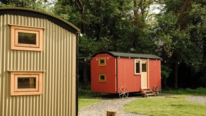 Two small, single-story cabins with corrugated metal exteriors, one beige and one red, situated in a wooded area with a gravel path leading to them. - Cosy en suite shepherd hut on the grounds of Historic building Samlesbury Hall (Samlesbury)