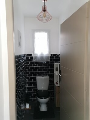 A compact bathroom with a black and white tiled wall, a white toilet, a small window with a white curtain, and a pendant light fixture. - New single-story house in the village center (Bédoin)