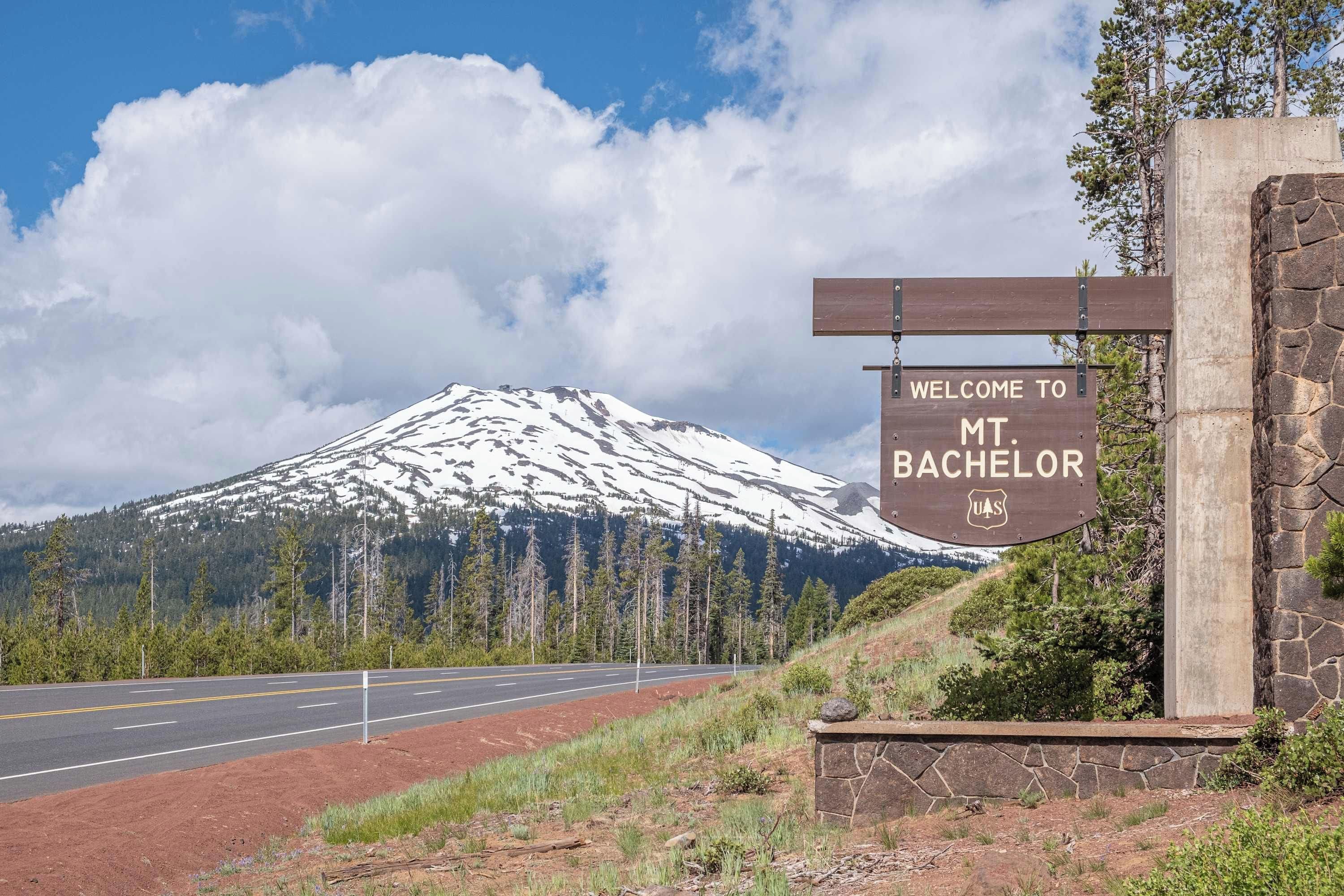 Una señal de tráfico da la bienvenida a visitantes a Mt. Bachelor, con una montaña nevada y cielo despejado al fondo.