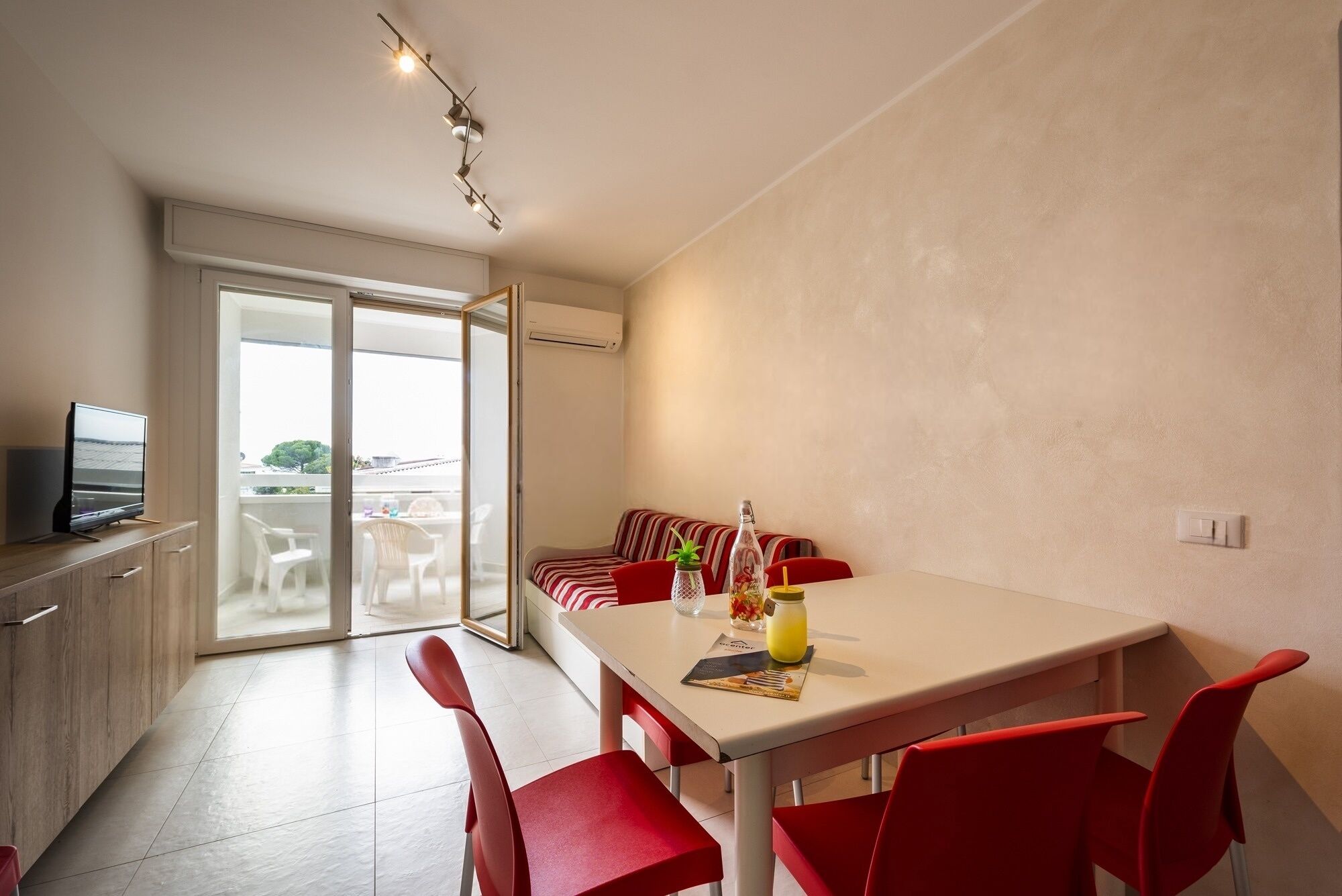 Un espace repas moderne avec une table blanche et des chaises rouges, un canapé avec des coussins à rayures et un téléviseur posé sur un meuble en bois.