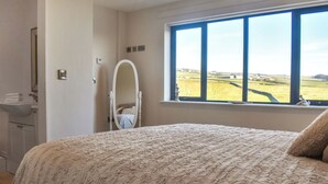 A bedroom with a large bed, a window with a view of the countryside, a bathroom with a sink and mirror, and a door. - The Wolfs Den - UK49083 (Laneshawbridge, near Colne)