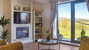 A living room with a flat-screen TV, bookshelves, a coffee table, and a large window with a view of the countryside. - The Wolfs Den - UK49083 (Laneshawbridge, near Colne)