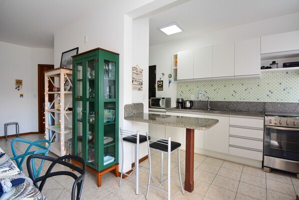 A kitchen with white cabinets, a green cabinet, a microwave, a coffee maker, and a stove. There are two bar stools and a dining table with chairs.