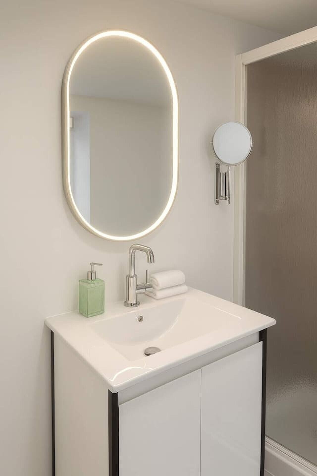 Une salle de bain moderne avec un lavabo blanc, un miroir doté d’un cadre lumineux, un distributeur de savon et un porte-serviettes.