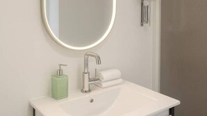 A modern bathroom with a white sink, a mirror with a lighted frame, a soap dispenser, and a towel rack.