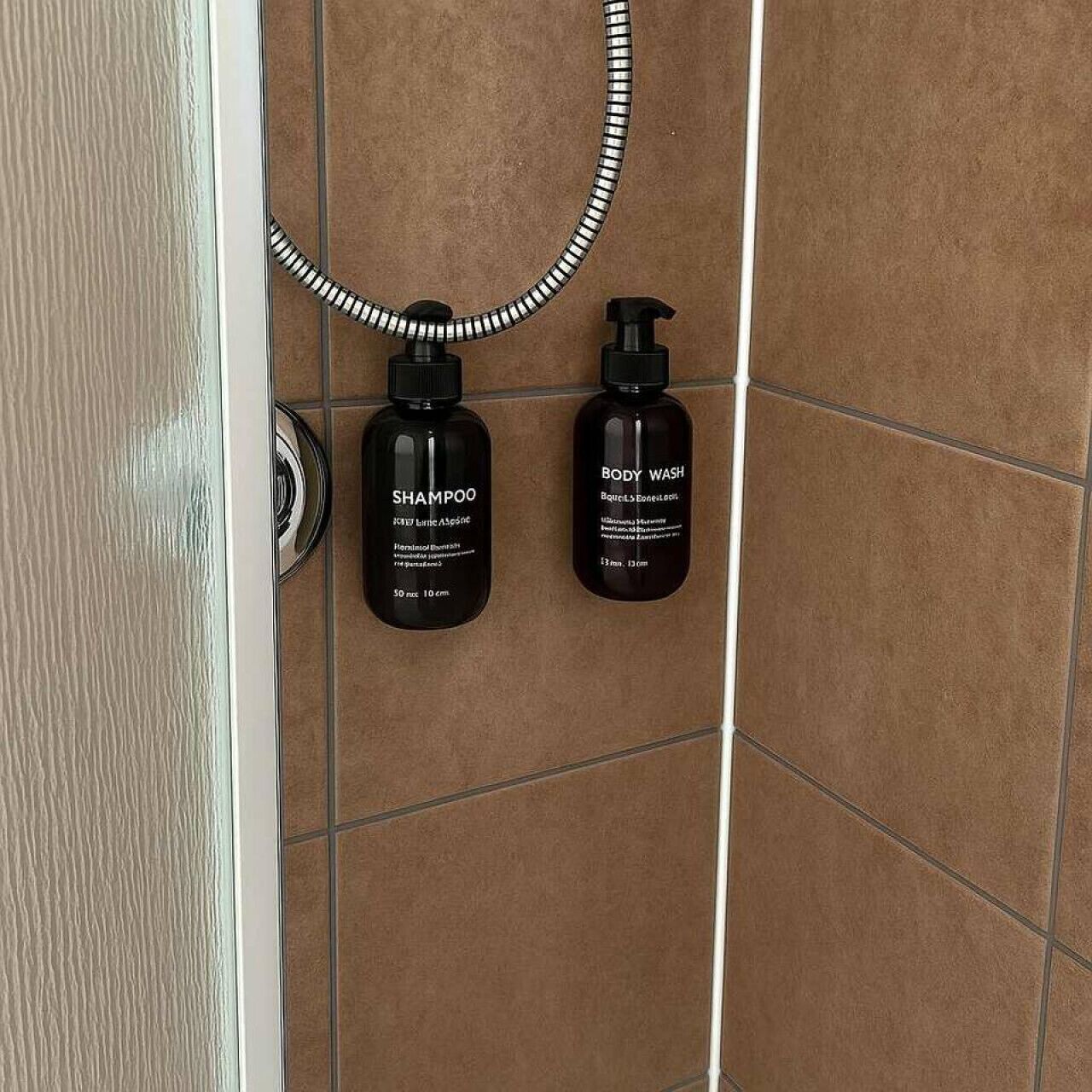 Une salle de bain avec deux bouteilles noires étiquetées « SHAMPOO » et « GEL DOUCHE » posées sur un mur carrelé.