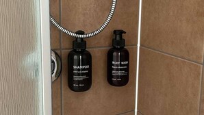 A bathroom with brown tiles, a shower, and two black bottles labeled "SHAMPOO" and "BODY WASH".