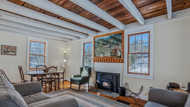 A cozy living room with a fireplace, wooden beams, and large windows. - Riverside Cottage on the Ottauquechee River (Woodstock)