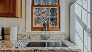 A kitchen with a granite countertop, a stainless steel sink, a window with a view of trees, and wooden cabinets. - Riverside Cottage on the Ottauquechee River (Woodstock)