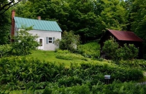 Une maison blanche avec un toit vert, entourée d’une végétation luxuriante et d  ’une petite construction dont le toit est rouge.