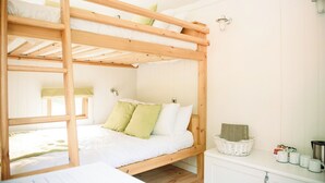 A bunk bed with a wooden frame, a white mattress, and green pillows. There is a white wicker basket on the nightstand, a white cabinet with a kettle, and a window with a curtain.