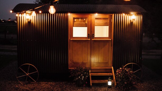 A small, single-room structure with a wooden door and windows, surrounded by a metal fence, and illuminated by string lights.