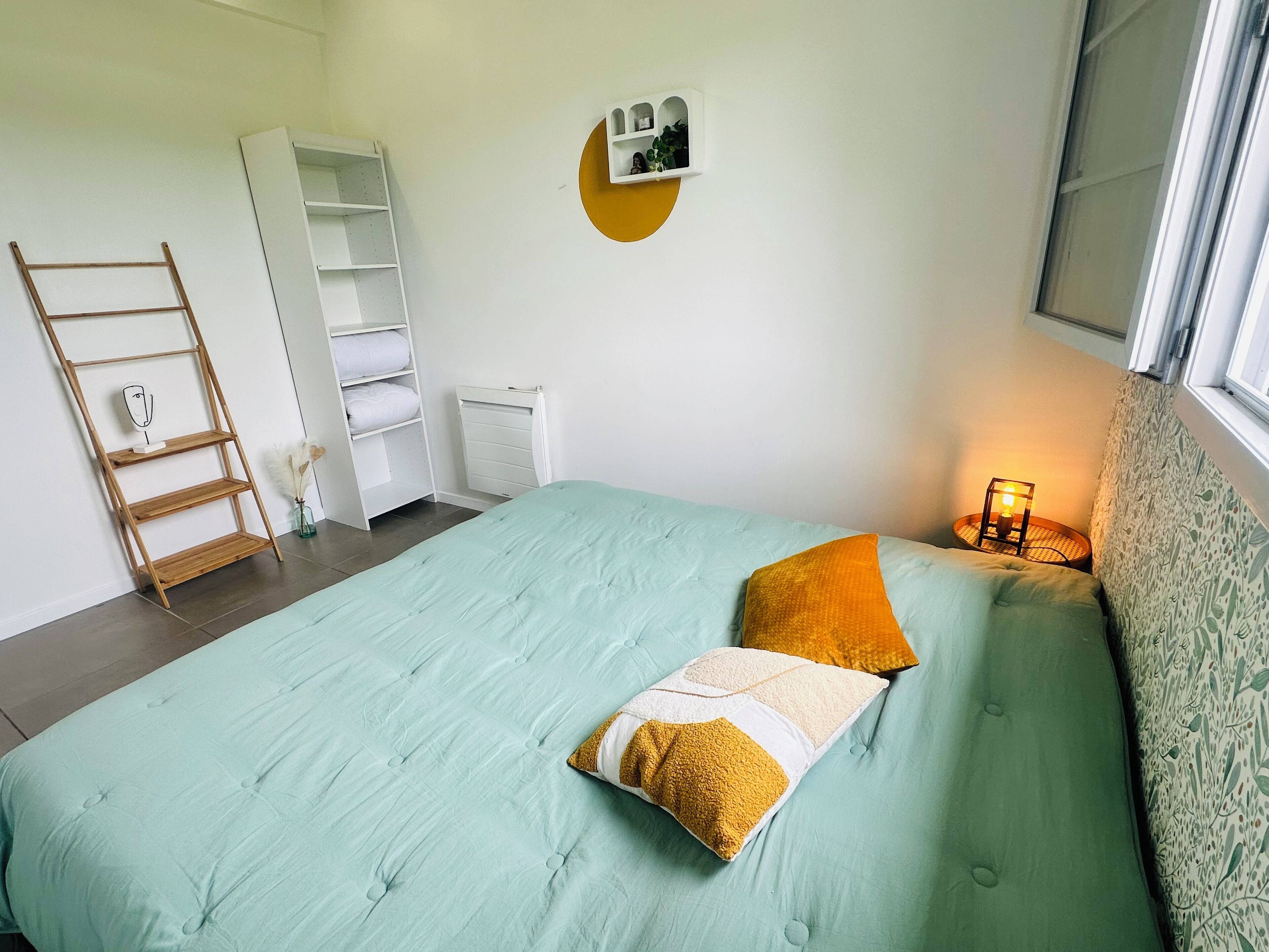Een slaapkamer met een bed, een ladder, een witte inbouwkast, een plank met planten en een raam met uitzicht.