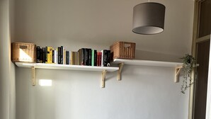 Una habitación con pared blanca, una estantería con libros y cestas, un escritorio gris, una silla de mimbre y una planta colgante.