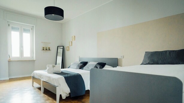 Una habitación de hotel moderna con una cama grande, una cama más pequeña, una mesita de noche con lámpara y una ventana con persianas.