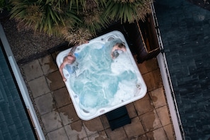 A hot tub with two people, surrounded by plants and a tiled patio. - Beach 600m • Sleeps 8 • Hot Tub • Pool • Dogs x2 (Holcombe)