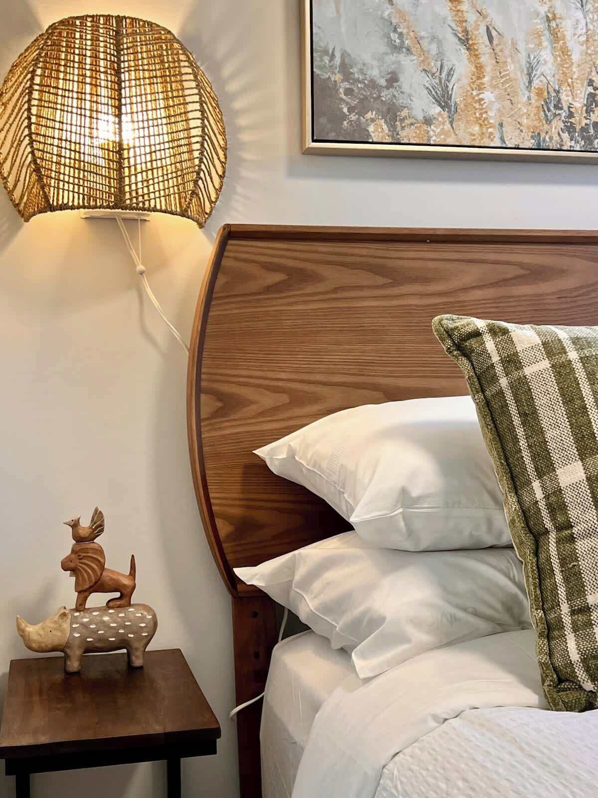 A neatly made bed with white pillows and a green and white patterned pillow, a small wooden nightstand with a decorative animal figurine, and a wall-mounted lamp with a woven shade.