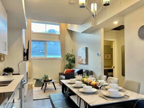A modern living room with a dining area, a sofa, a coffee table, and a large window. - Brand new - 3 Bedroom Sky Ridge End Unit Condo (Harvie Heights)