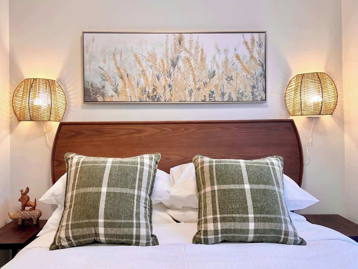 Una cama con cabecera de madera, dos almohadas a cuadros, una colcha blanca y dos lámparas fijadas a la pared con un diseño tejido.