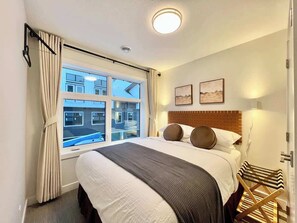A hotel room with a large bed, a window with curtains, a wooden headboard, and a view of buildings outside. - Brand new - 3 Bedroom Sky Ridge End Unit Condo (Harvie Heights)