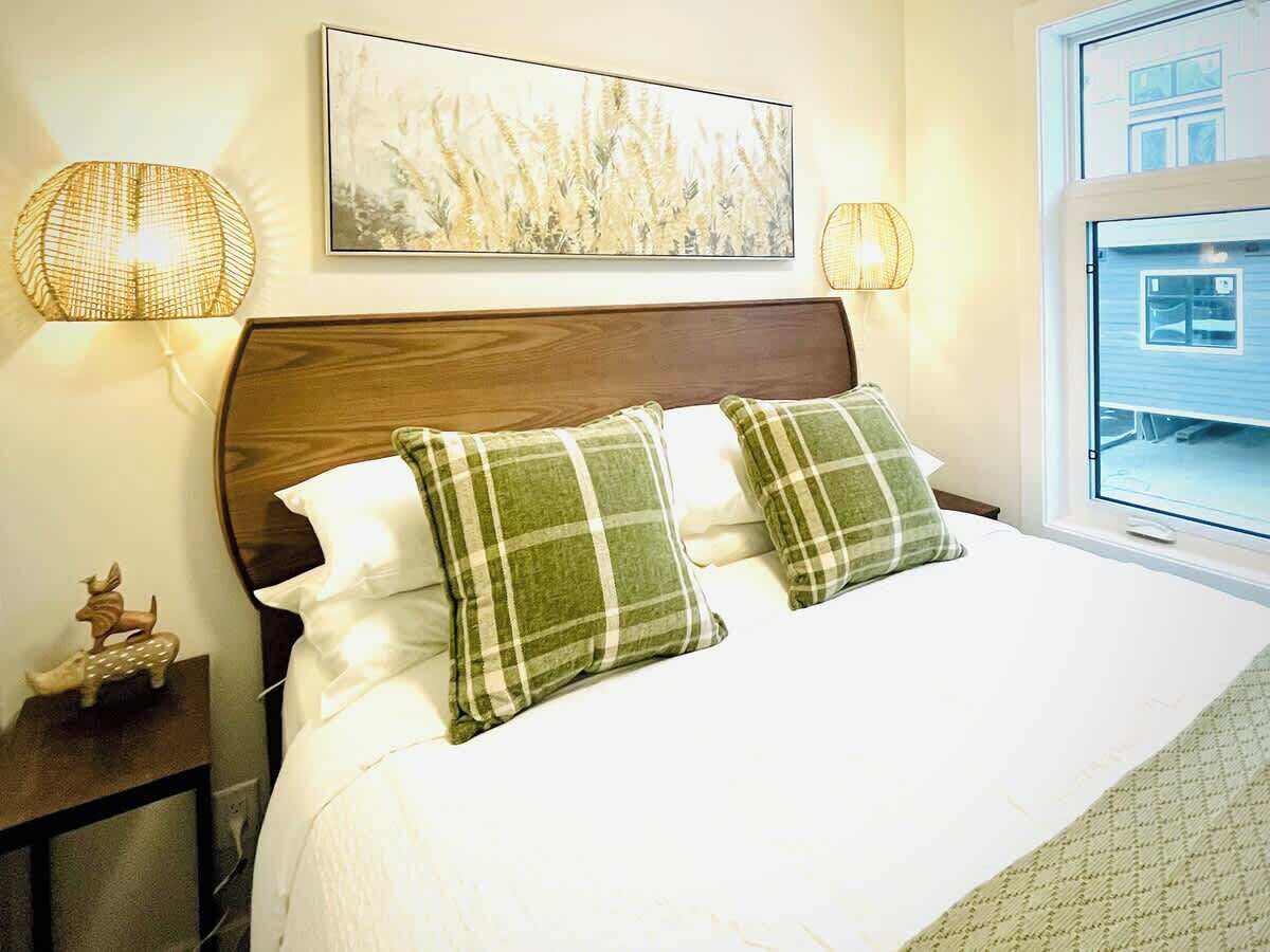 A neatly made bed with a wooden headboard, two green and white checkered pillows, and a white comforter. There is a framed artwork above the bed, two wall-mounted lamps on either side, and a window with a view of a building.