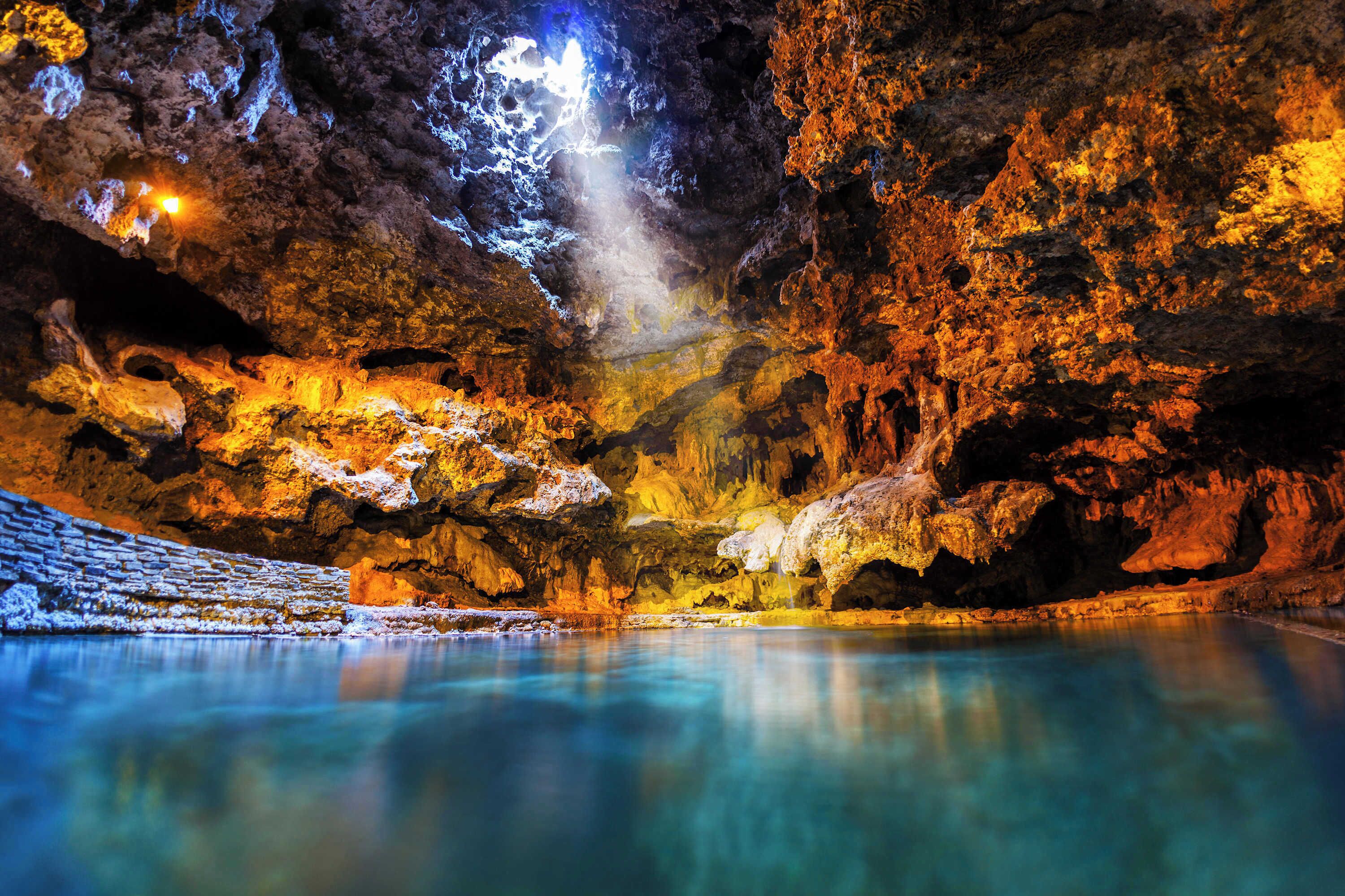 En grotta med stalaktiter och stalagmiter, en vattenpöl och en ljuskälla som lyser upp grottan.