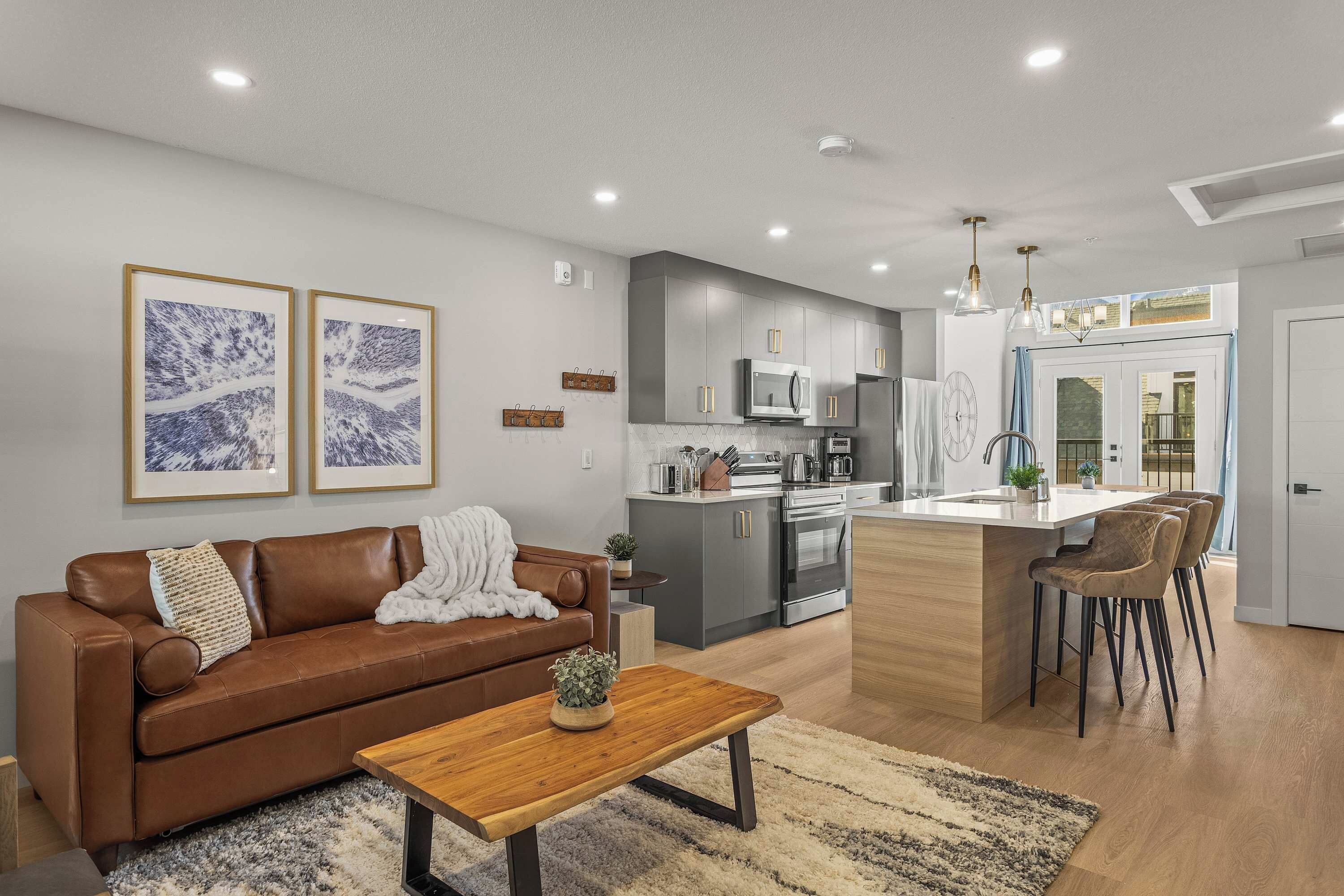 A modern living room with a brown leather sofa, a wooden coffee table, and a kitchen area featuring grey cabinets, stainless steel appliances, and a central island with bar stools.