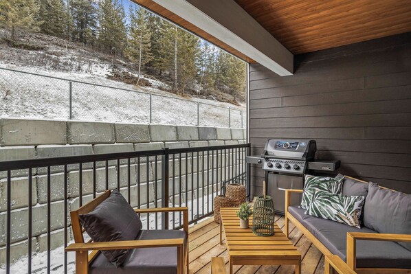A covered outdoor seating area with a grill, wooden furniture, and a view of a snowy landscape.