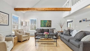 A spacious living room with a large gray sofa, a beige armchair, and a glass coffee table. There are two windows with white frames, a wooden ceiling beam, and a colorful abstract painting on the wall. - "Big Sky" Retreat Atop Sunapee / Ski + Lake Life (Newbury)