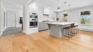 A modern kitchen with white cabinets, stainless steel appliances, a central island with stools, and large windows. - "Big Sky" Retreat Atop Sunapee / Ski + Lake Life (Newbury)