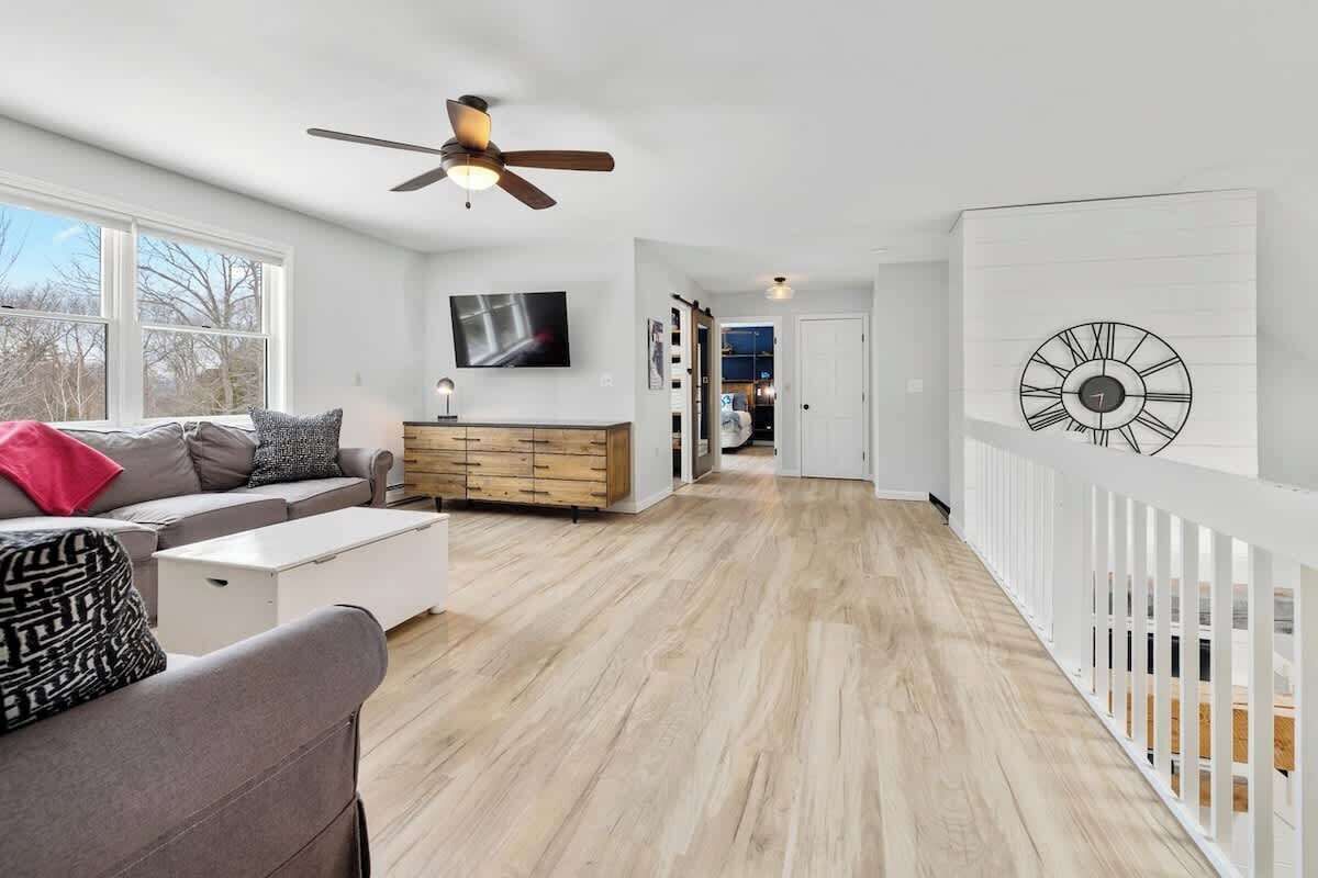Un salon moderne avec un canapé, une table basse et un meuble en bois. On y trouve un ventilateur de plafond, une horloge au mur et une vue sur l’extérieur par une fenêtre.