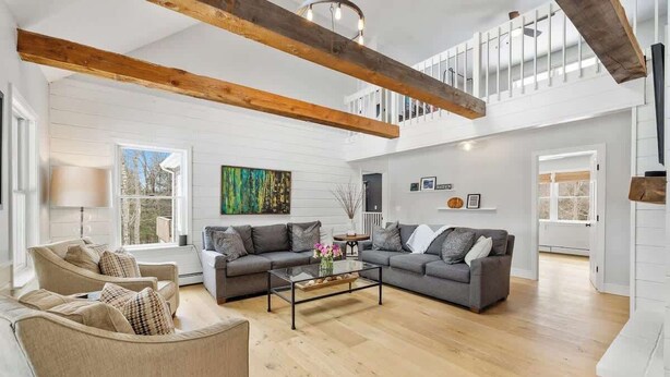 A spacious living room with a large sectional sofa, a glass coffee table, and a high ceiling with exposed wooden beams. - "Big Sky" Retreat Atop Sunapee / Ski + Lake Life (Newbury)