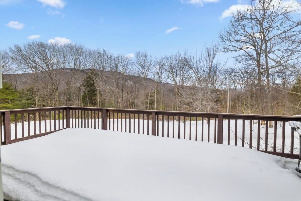 Ein mit Schnee bedeckter Balkon mit Holzgeländer, umgeben von kahlen Bäumen und einer hügeligen Landschaft im Hintergrund.