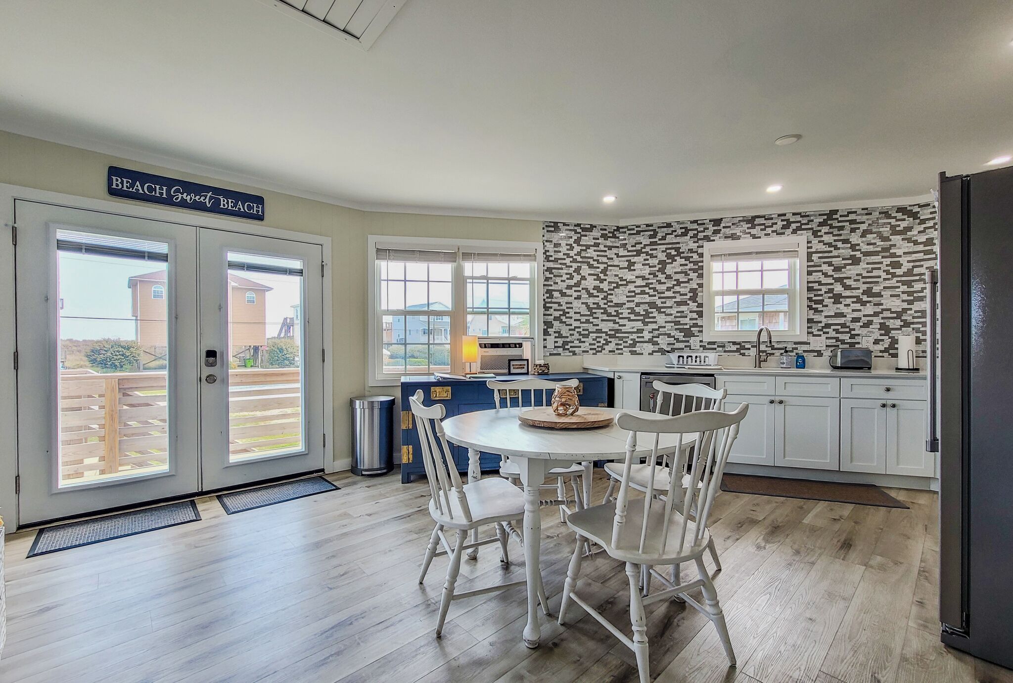 A kitchen with white cabinets, a dining table with chairs, a sign reading "Beach Sweet Beach", and a view of the outside.