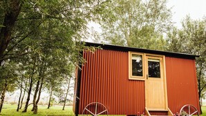 A small, single-story wooden structure with a door and windows, surrounded by trees and grass. - Cosy en suite shepherd hut on the grounds of Historic building Samlesbury Hall (Samlesbury)