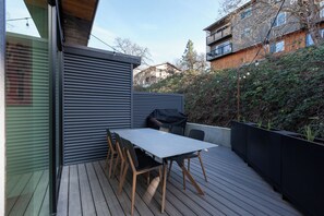 A wooden deck with a table and chairs, a grill, and a view of neighboring buildings.