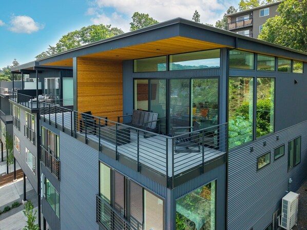 A modern multi-story building with large windows, a wooden deck, and a balcony with seating.