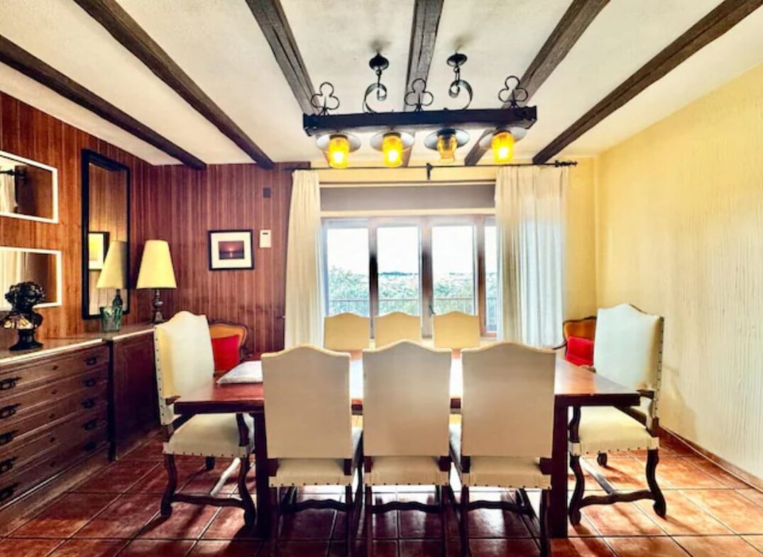 Un coin repas avec une table et des chaises en bois, une fenêtre avec des rideaux et un plafond avec des poutres apparentes et des luminaires.