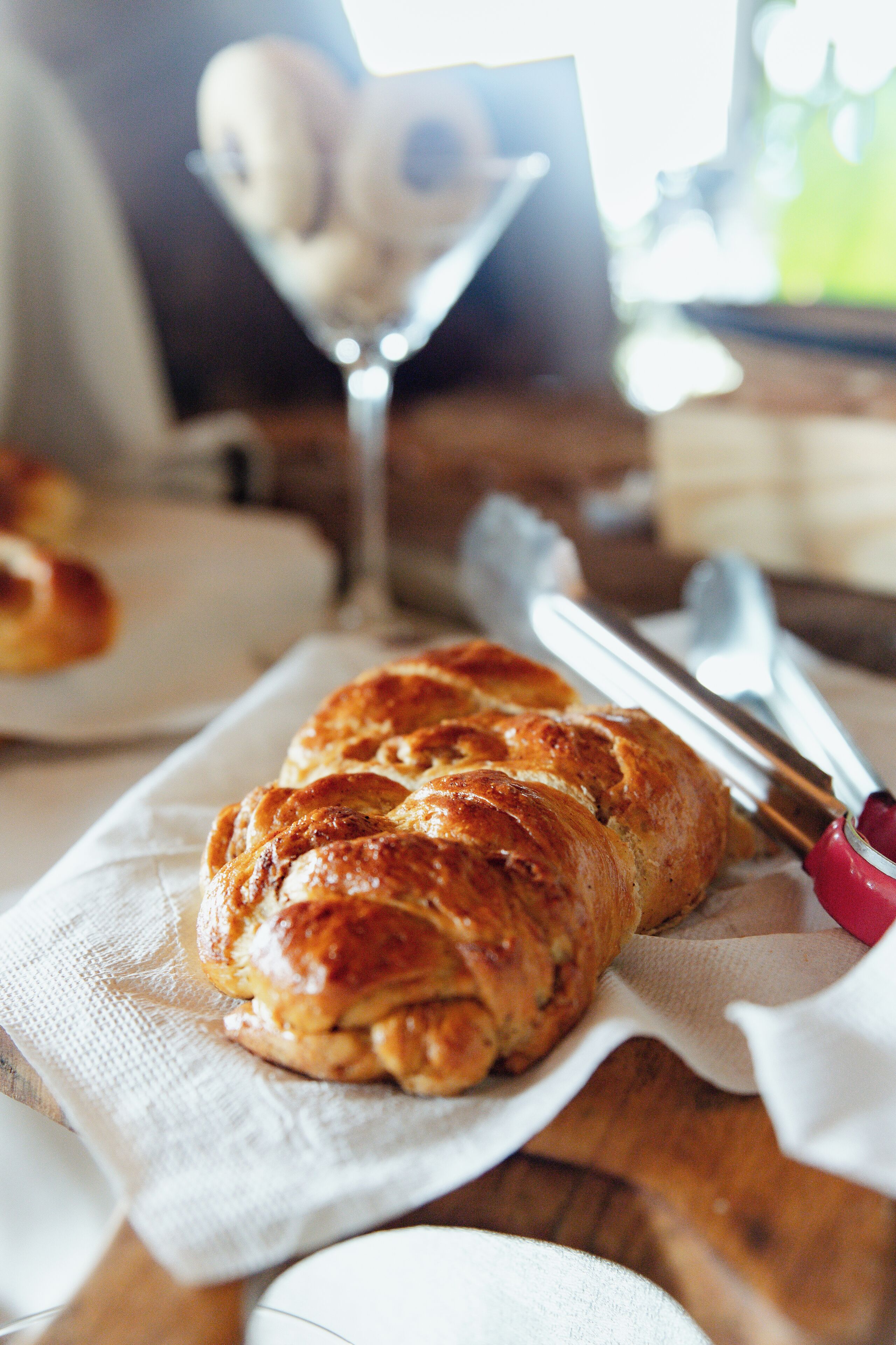A loaf of bread on a white napkin, a dessert in a glass, and a knife with a red handle on a wooden table.