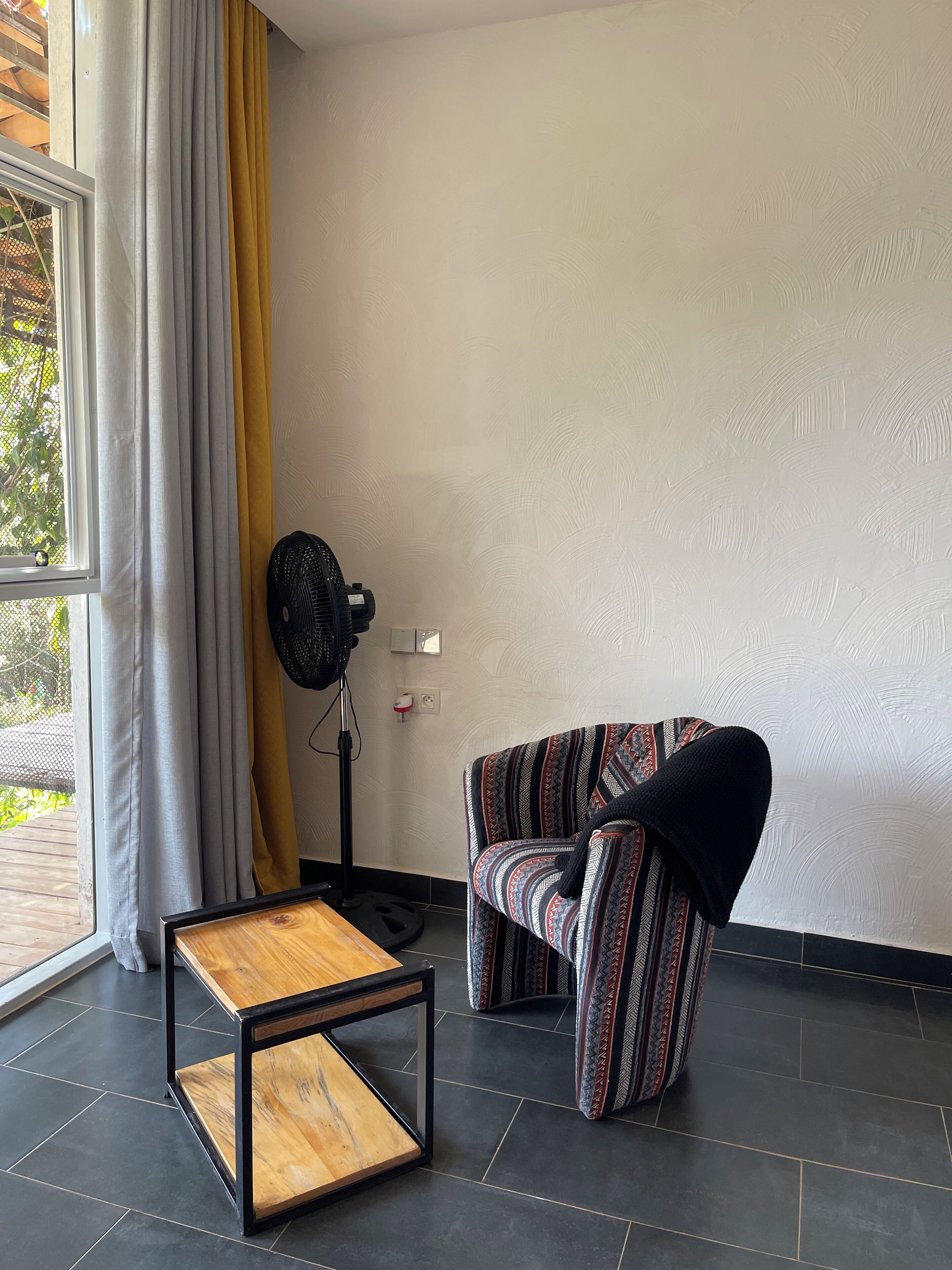 A modern living room with a wooden coffee table, a patterned sofa, a standing fan, and a large window with curtains.