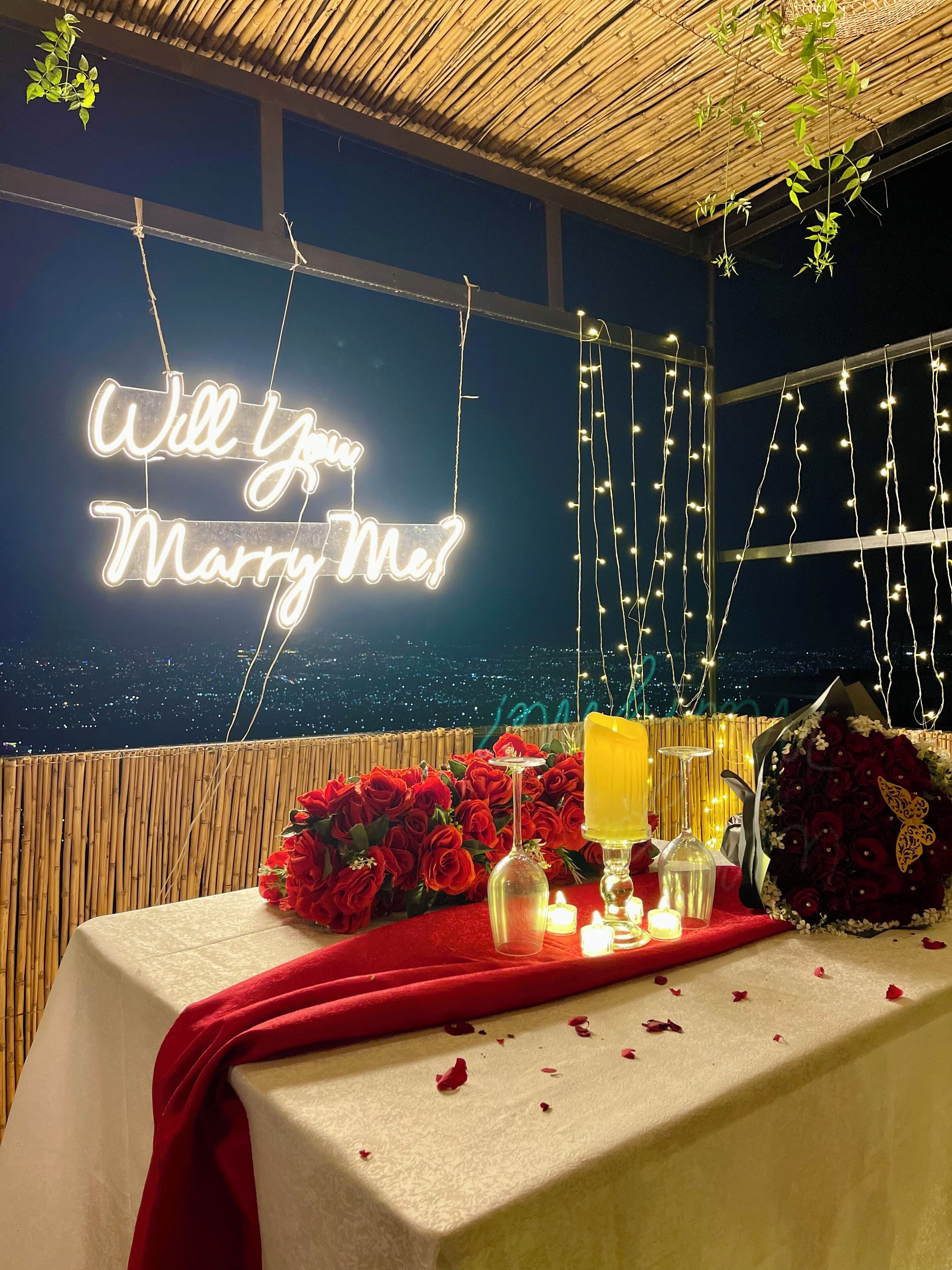 A romantic outdoor setting with a neon sign reading "Will you marry me?" and a table adorned with red roses and candles.